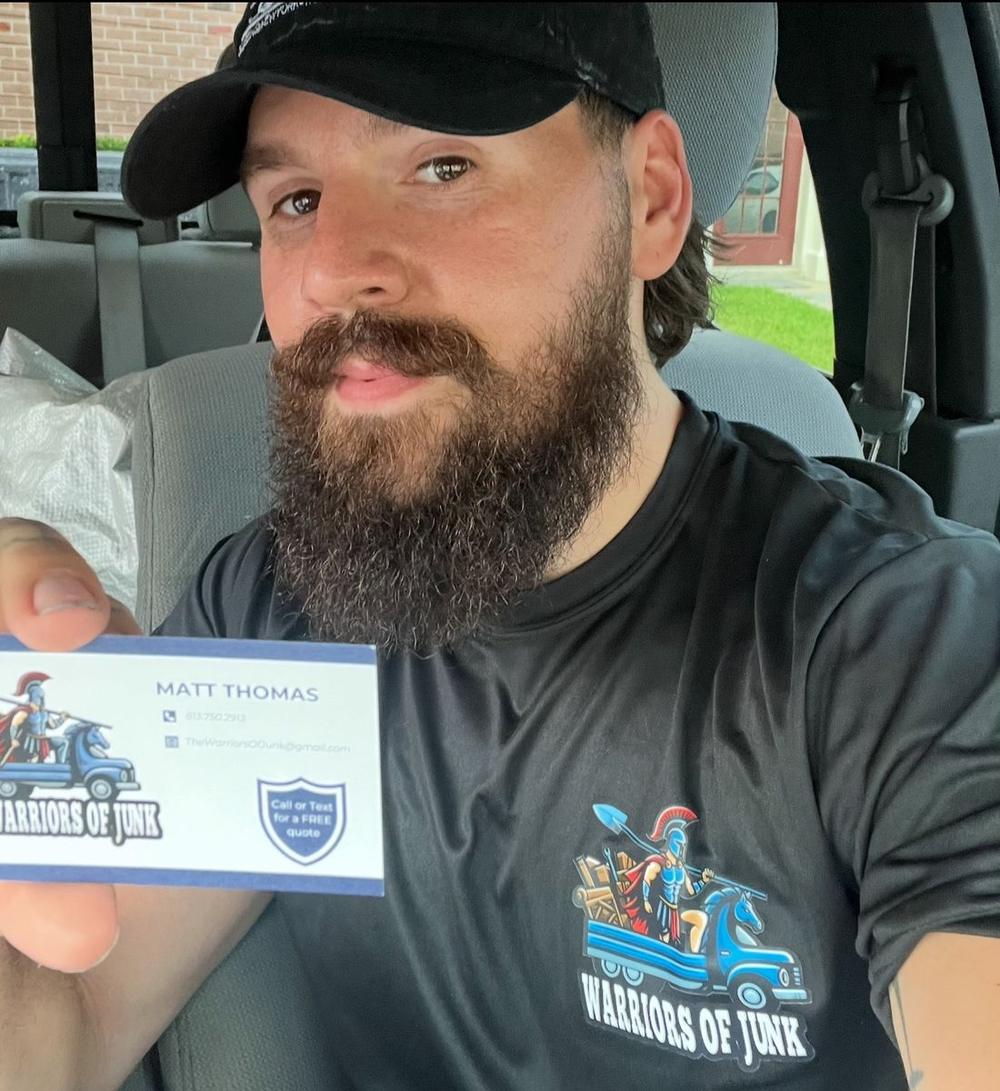 Matt Thomas in a branded Warriors of Junk shirt, holding up his own business card with the Spartan and truck logo printed on it.