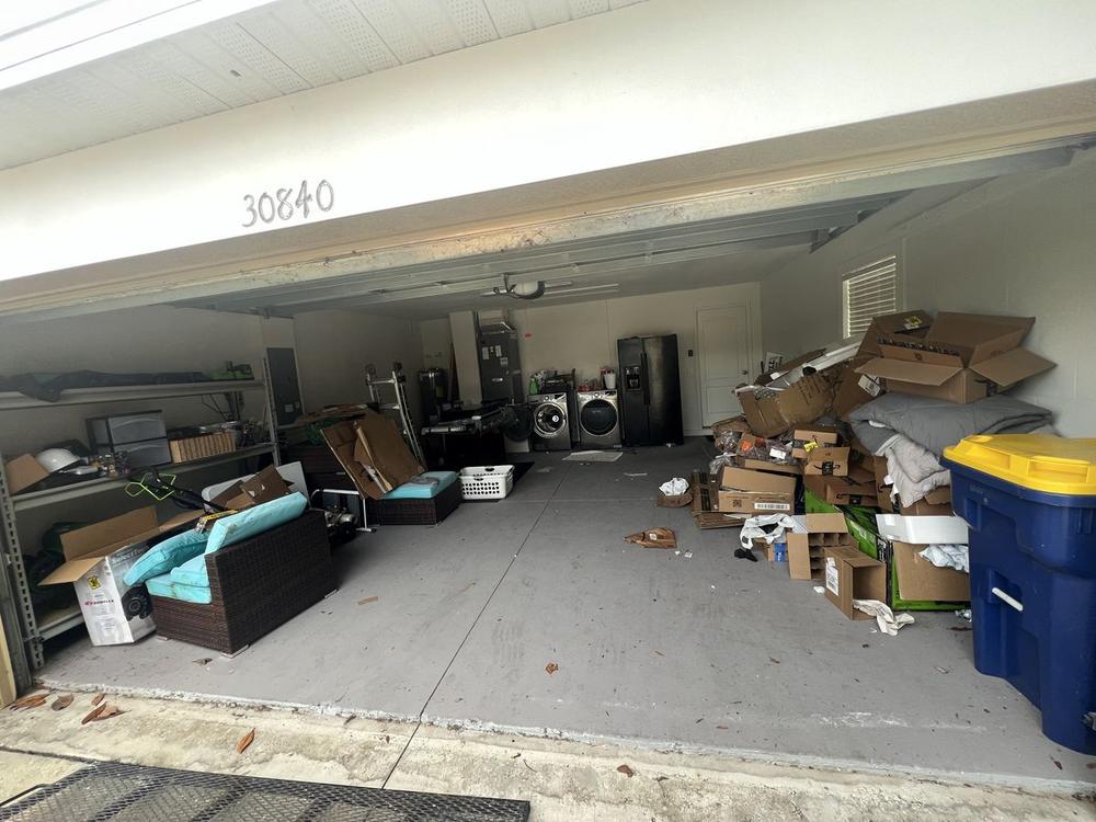 A two-car garage packed to the back wall with a sectional sofa, a stacked washer and dryer, flattened cardboard and loose debris across the floor. The house number 30840 is painted on the header beam.