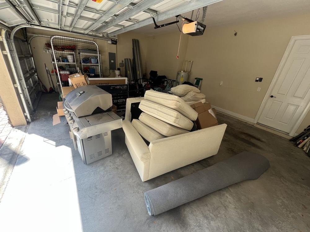 A beige garage with mattresses stacked on a sofa, a rolled carpet on the floor and boxes crowding the doorway, leaving no room to walk through.