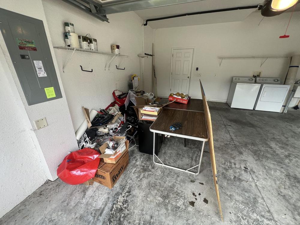 A garage beside a grey electrical panel, its floor taken up by a folding table, stacked boxes, a red bag and loose household clutter.