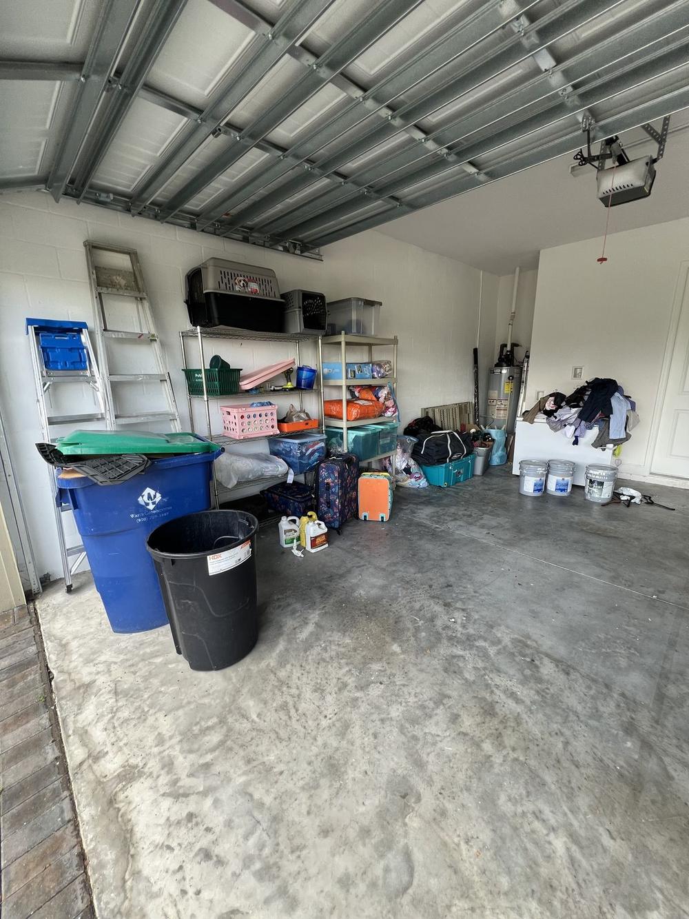 The same truss-roofed garage with the floor emptied, only a blue recycling bin and a black trash can remaining beside the shelving.
