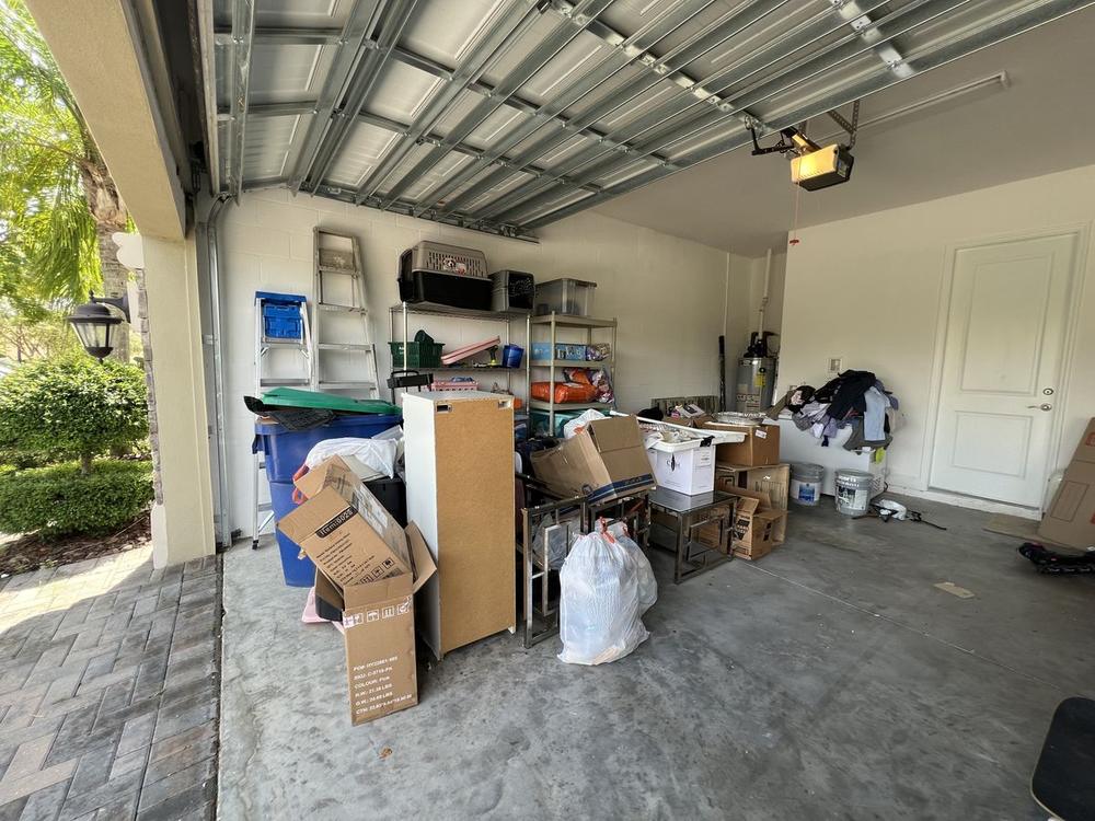 A garage under exposed metal roof trusses, with a green folding table, a wooden cabinet, broken-down cardboard and full white bags covering the floor.