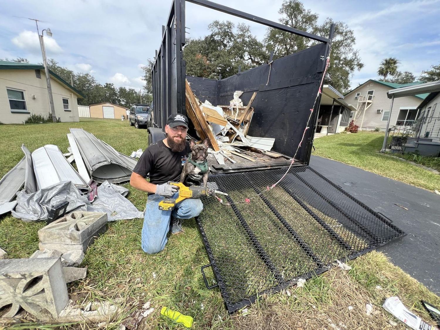 Matt kneeling on the grass in a branded black shirt and cap with his French bulldog, a reciprocating saw in his gloved hand, beside his black mesh-gate dump trailer loaded with broken framing lumber and white painted trim.