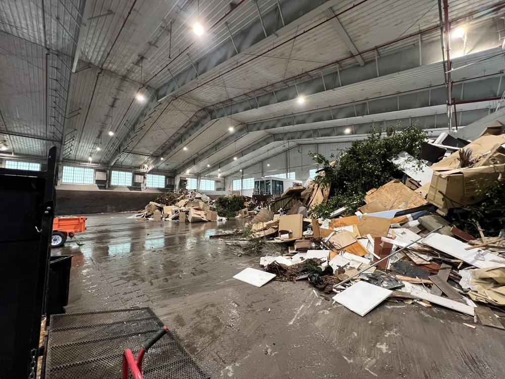 A wide commercial warehouse interior with broken pallets, timber and packaging debris spread across the concrete floor under the roof lights.