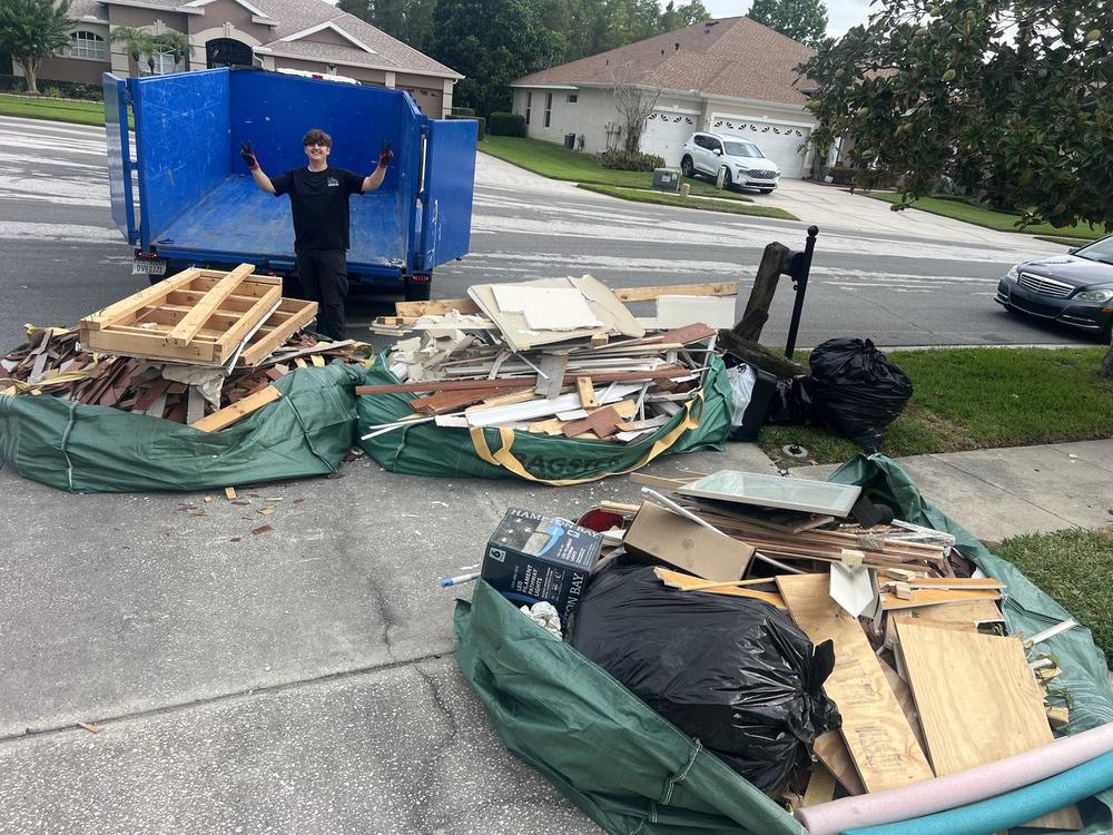 Matt standing in the blue dump trailer with his arms raised over a driveway spread of pallets, broken lumber, tarps and filled contractor bags outside a house.