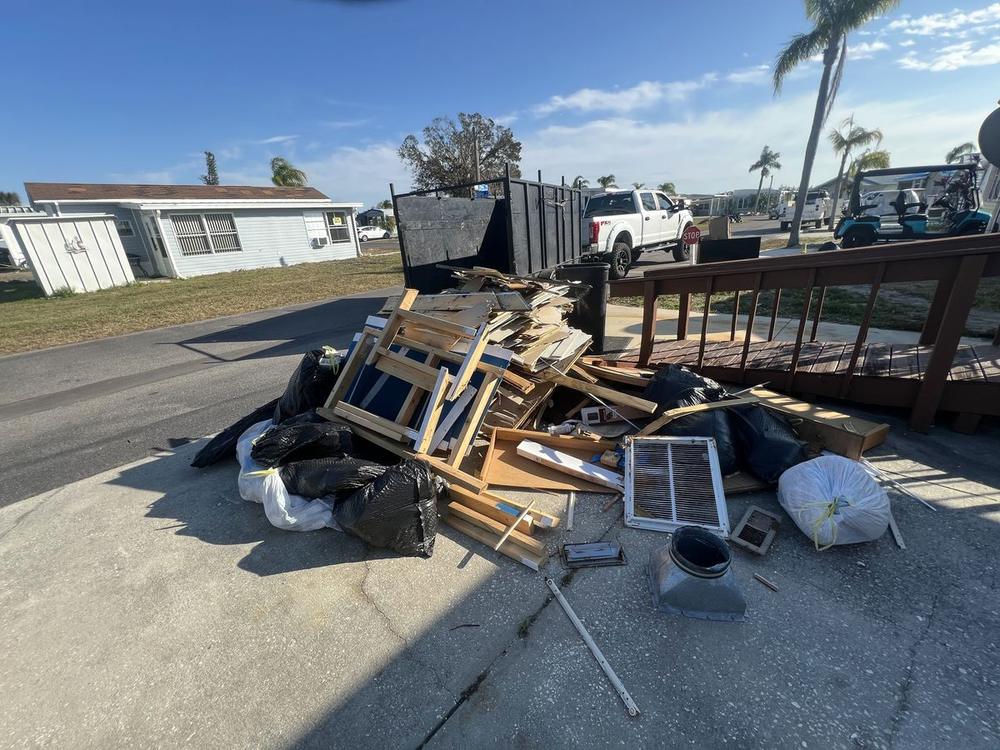 A driveway pile waiting for pickup: black contractor bags, plastic-wrapped bundles, a raw pine cabinet carcass, a white return-air grille and broken lumber, with the loaded trailer and a white pickup behind it.