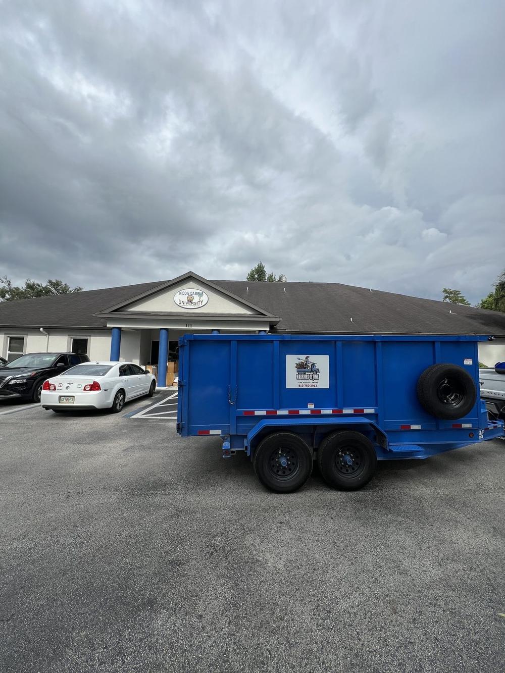 The blue dump trailer with the Warriors of Junk logo panel on its side, parked and ready outside a low building under a bank of grey cloud.